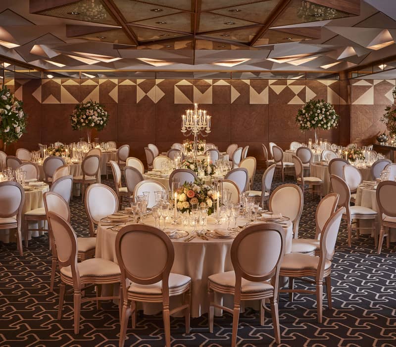 A luxurious ballroom with elegant tables, white chairs, floral centerpieces, candelabras, and warm, modern decor.