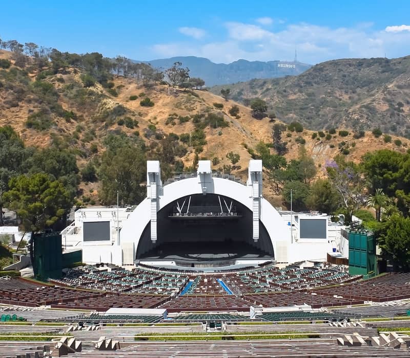Outdoor amphitheatre with empty seats, central stage, and Hollywood Hills in background under blue sky; Hollywood sign visible.