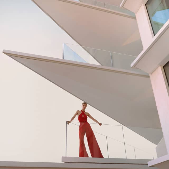 A tall female model with brown hair poses in a striking red jumpsuit on a glass window balcony that is located on a pointed edge of an architectural building