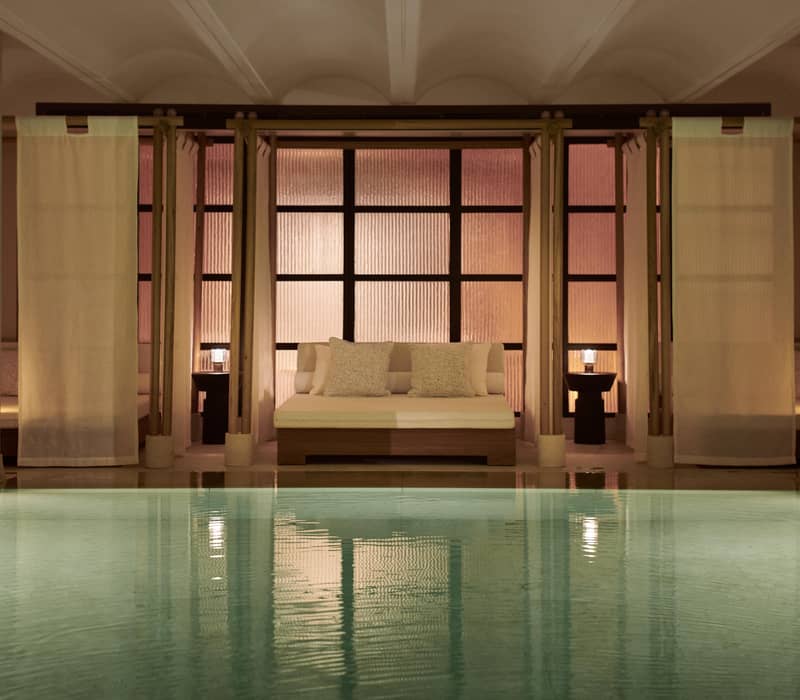 Luxurious indoor pool at Claridge's Spa with serene lighting, elegant daybeds draped with curtains, and soft, neutral tones for ultimate relaxation.