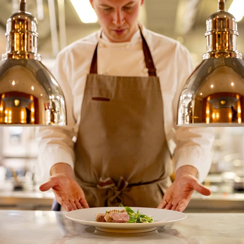 Chef in white uniform and apron presenting a plated gourmet dish under warm pendant lights in a professional kitchen setting.
