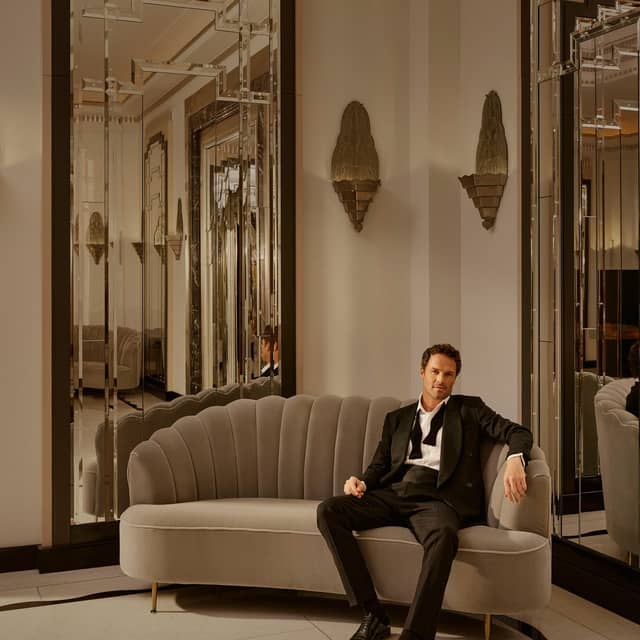 A man in a dinner suit sits casually on a grey, scalloped sofa in an opulent, art deco-inspired room.
