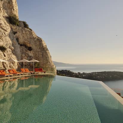 Cliffside infinity pool with orange loungers and parasols overlooking the Mediterranean Sea and coastline under a clear sky.