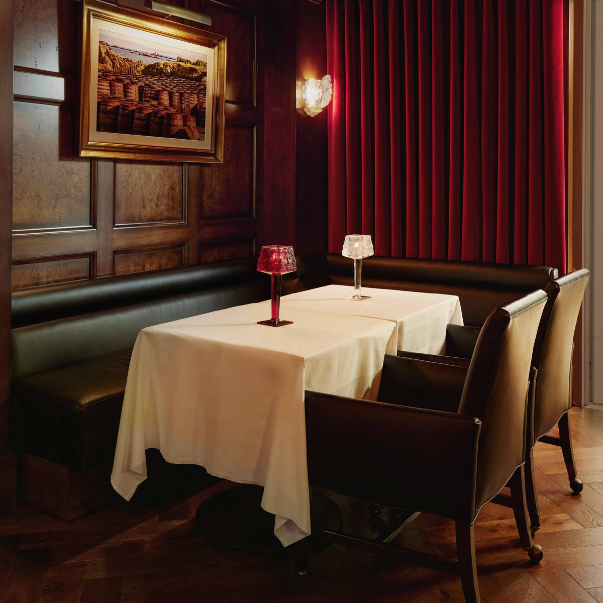 Intimate lounge setting with leather seating, a white tablecloth, crystal lamps, red velvet curtains, and rich wood paneling.