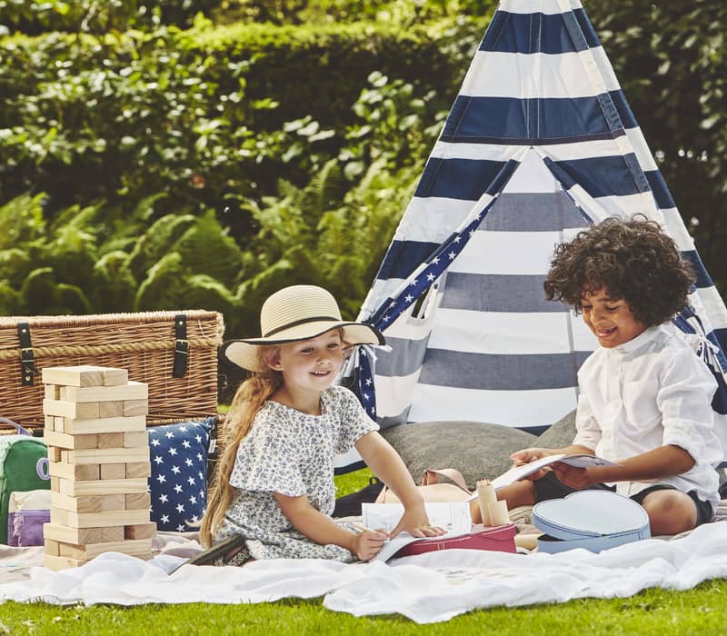 Children enjoying an outdoor picnic with games, a striped tent, and wicker baskets, set in a lush green garden.