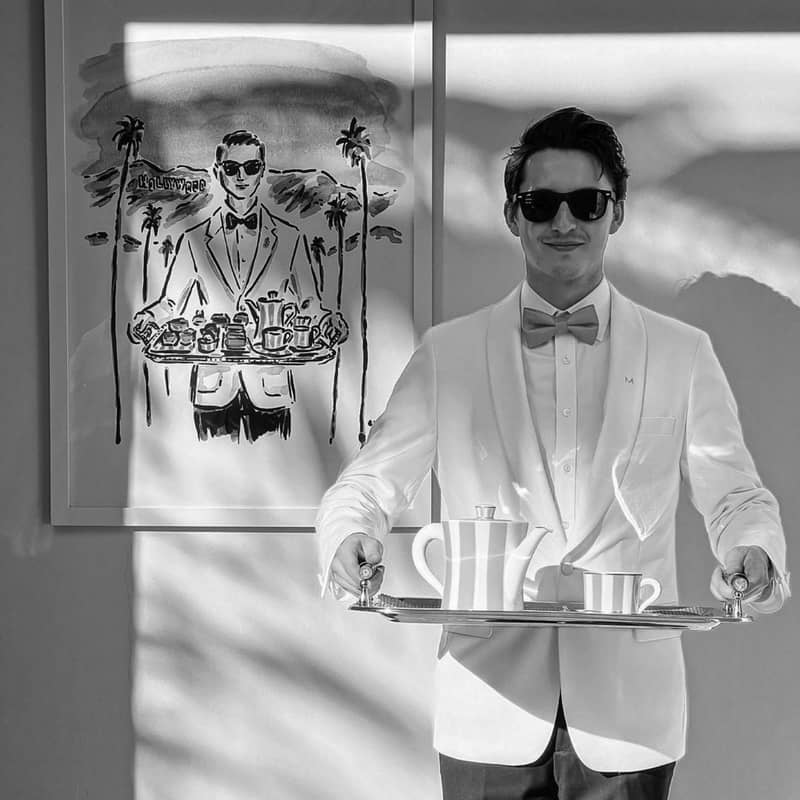 A waiter in a white tuxedo and sunglasses carries a silver tray with tea, mirroring a stylish illustration behind him.