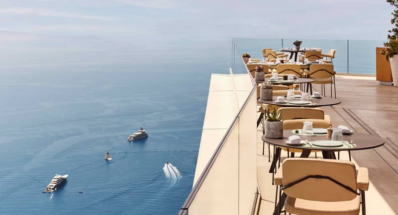 Clifftop restaurant terrace with neatly set tables and beige chairs overlooking a panoramic view of the Mediterranean Sea, with yachts and boats visible below on the water.