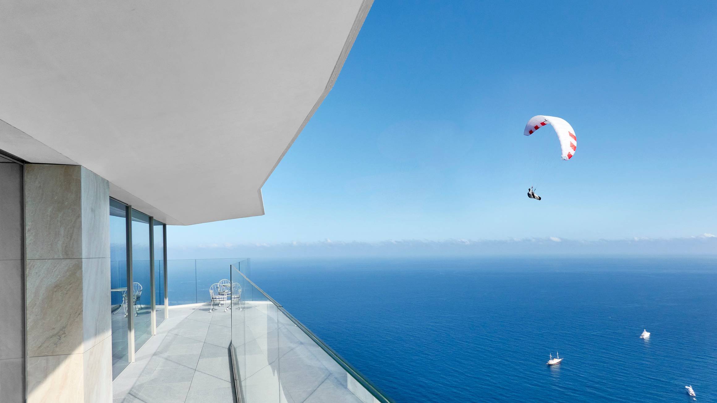 A modern terrace overlooking a vast blue ocean with yachts and a paraglider soaring under a clear sky.