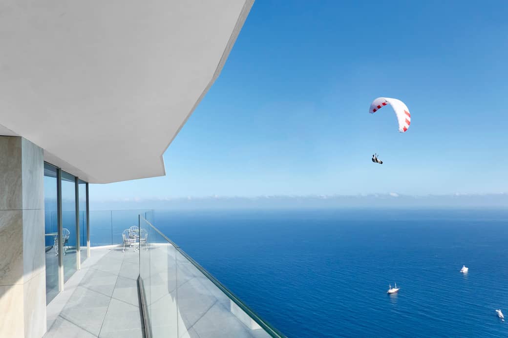 A modern terrace overlooking a vast blue ocean with yachts and a paraglider soaring under a clear sky.
