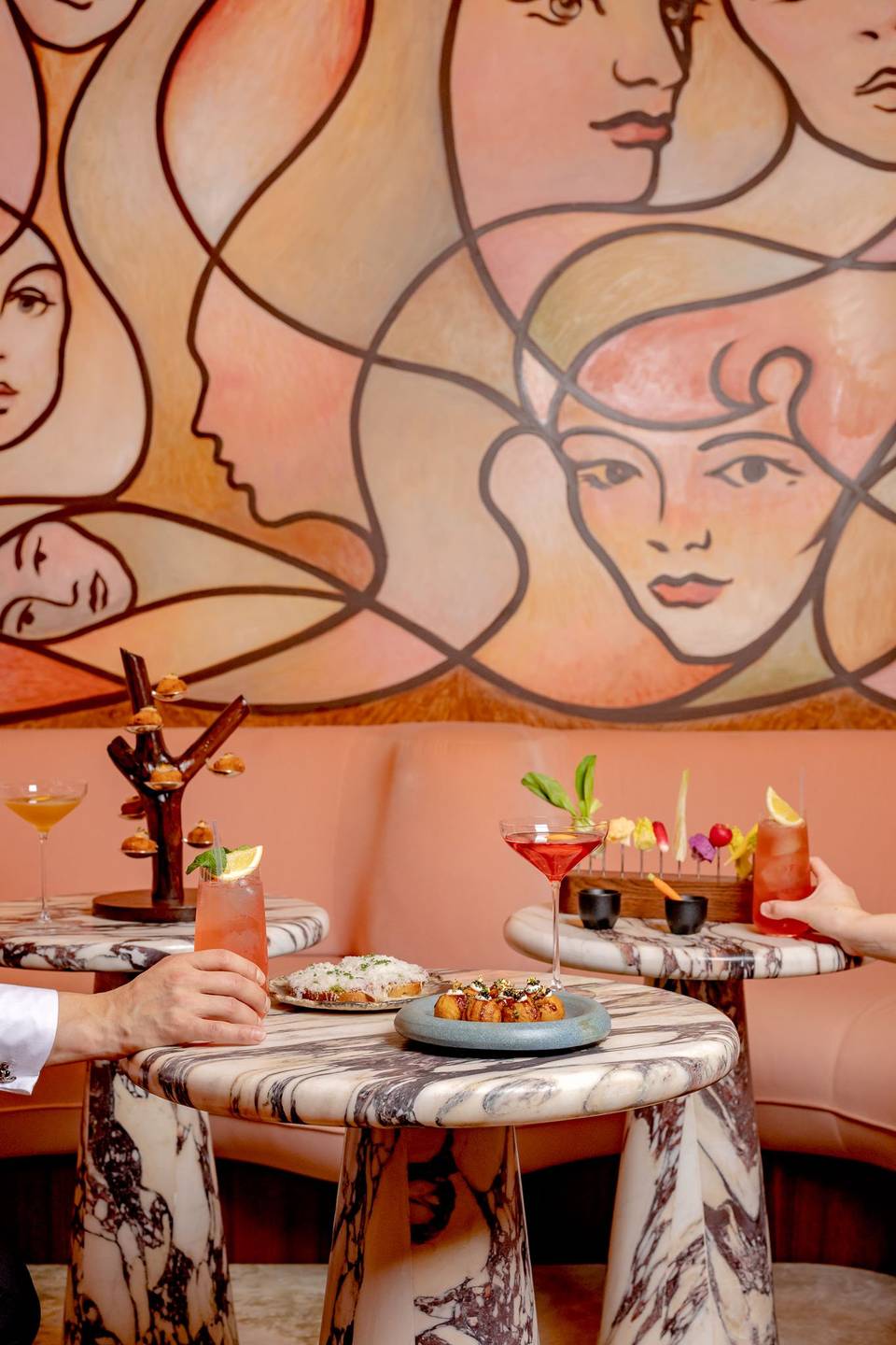 Artful cocktails and small plates on marble tables, set against a peach-toned mural with abstract feminine faces.