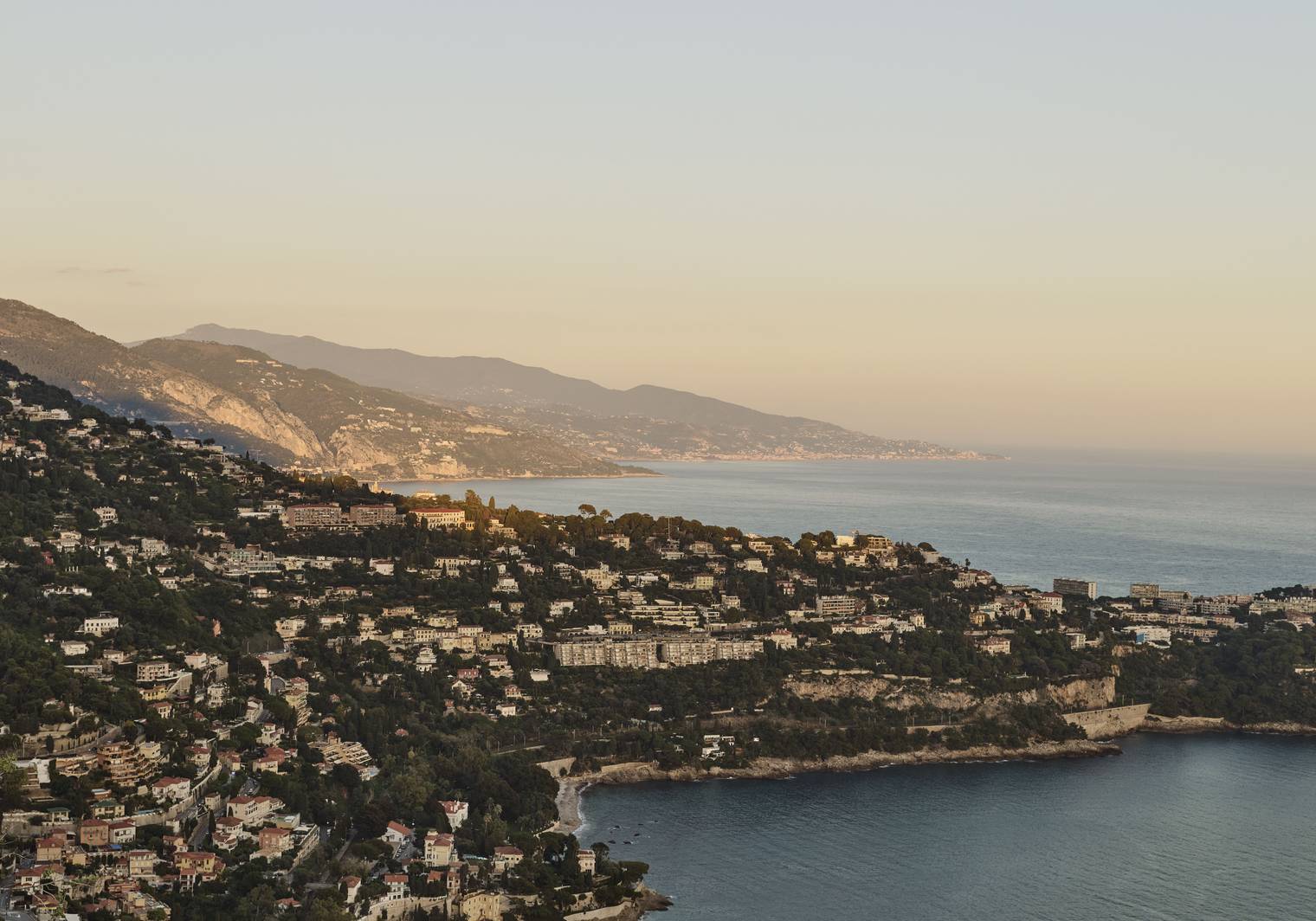 Aerial view of the French Riviera coastline at sunset, with hillside villas, lush greenery, and the Mediterranean Sea.
