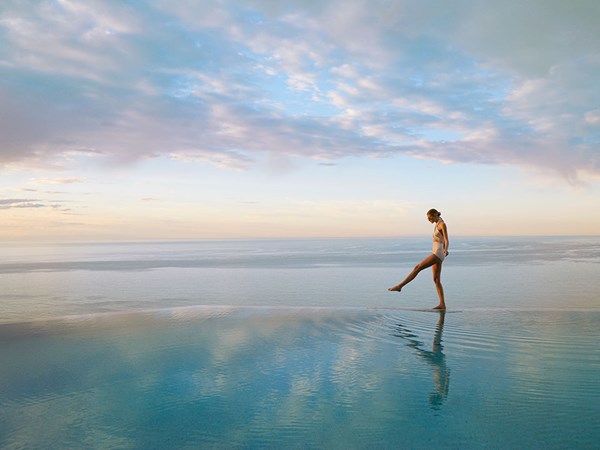 A lady in a swimsuit walks along the infinity pool edge infront of the sea and a beautiful sunset