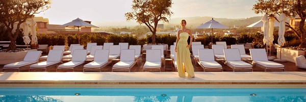 A woman in a flowy yellow outfit stands infront of white deckchairs and a blue pool