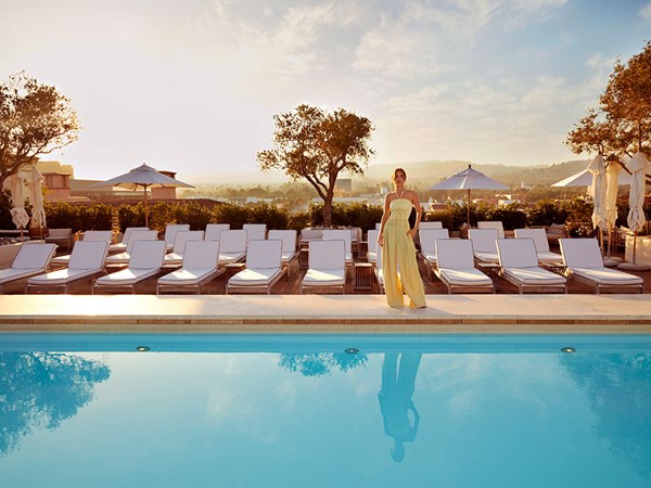 A woman in a flowy yellow outfit stands infront of white deckchairs and a blue pool