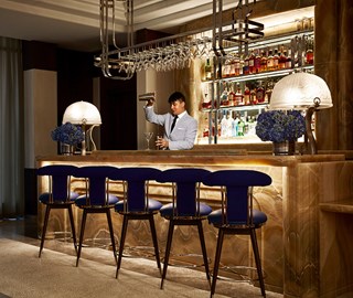 The Maybourne Bar at The Maybourne Beverly Hills. A bartender pours a cocktail behind a bar. There are five stools at the bar. Flowers and lamps are placed either end of bar.