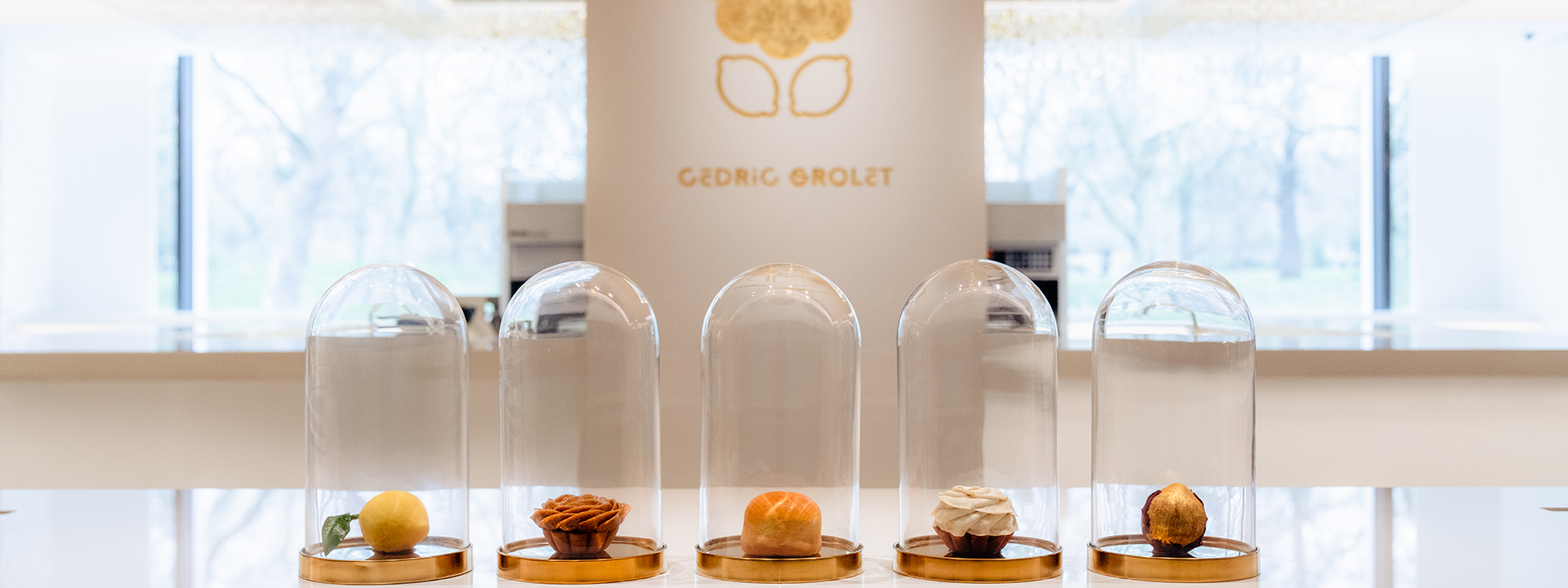 Image shows five cakes in cloches on a counter top. In the background a gold Cedric Grolet counter is visible.