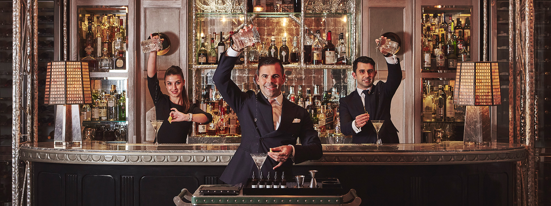 The Connaught Bar. The image shows three bar tenders - two men and one woman - who are making and pouring cocktails. They are all smiling and looking at the camera. They are all standing in front of a well-stocked bar. 