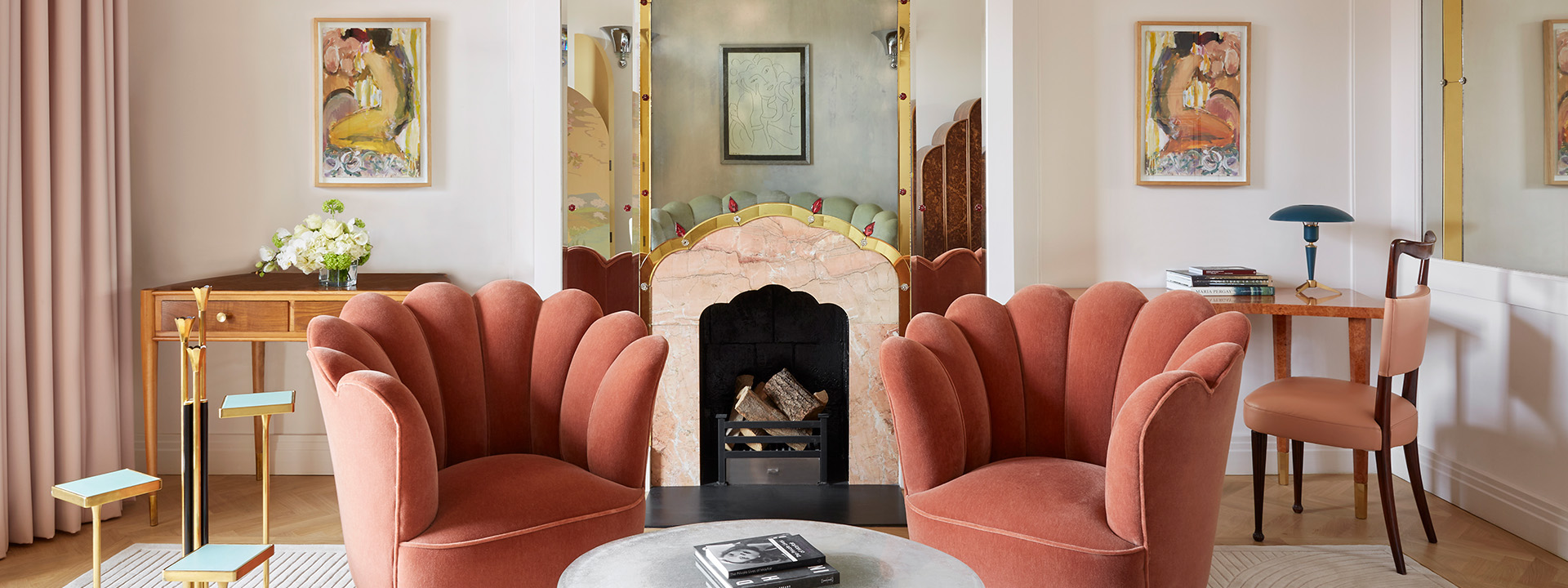 The Mayfair Terrace Suite. An image of a room with two blush coloured chairs with scalloped shape backs. The chairs are arranged between a table, in the background is a mirror and ornate wall. 