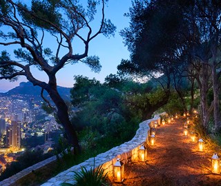 The Maybourne Riviera. This image is taken at dusk, and shows the town to the left of the image and a pathway lit by candles on the right.
