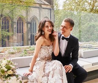 Weddings at The Berkeley. This image shows a man and woman on their wedding day. They are sitting inside smiling and laughing, behind them is a church and the bride's bouquet is on the left of the image.